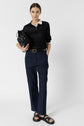A person stands against a plain background, wearing a black polo shirt, a pantalon coton bleu marine coupe droite, black sandals, and holding a black bag under one arm.
