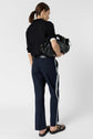Person with brown hair in a ponytail, wearing a black top and a pantalon coton bleu marine à coupe droite with white bandes latérales, and black sandals, holding a large black handbag, standing against a plain white background.