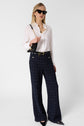 Woman wearing a white blouse, navy plaid high-waisted wide-leg trousers, black pointed shoes, black sunglasses, and carrying a small black purse with a gold chain strap. She stands against a plain white background.