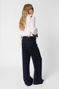 A woman with long blonde hair, wearing black sunglasses, a light pink blouse, and navy blue checkered wide-leg pants, stands facing away, holding a black handbag over her shoulder against a white background.