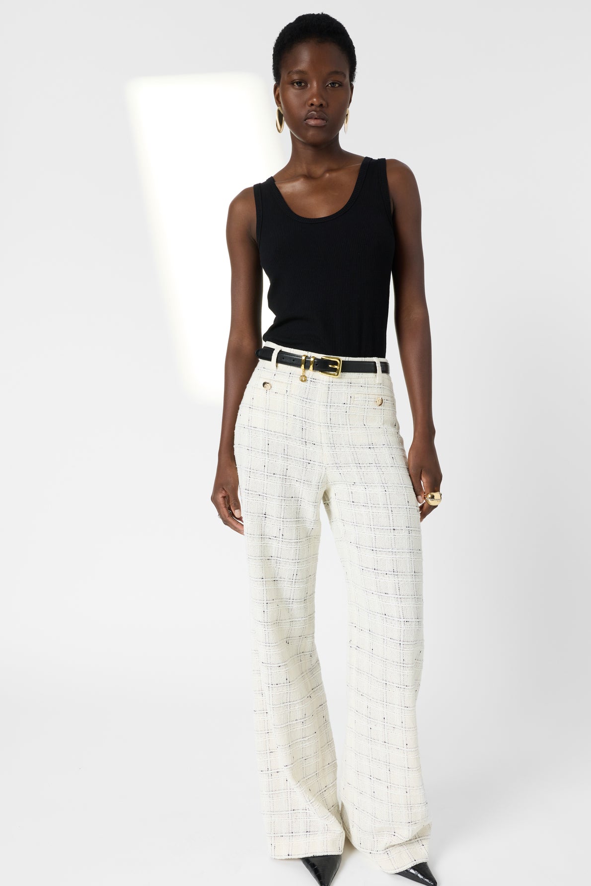 A woman stands against a white background wearing a black sleeveless top, high-waisted white plaid pantalon large trousers with a black belt, and black pointed shoes. She has short hair and wears hoop earrings and rings, effortlessly embodying the timeless elegance of Gerard Darel.