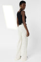 Woman standing and looking over her shoulder, wearing a sleeveless black top, wide-leg white plaid pantalon large from Gerard Darel paired with a black belt and gold hoop earrings against a white background.