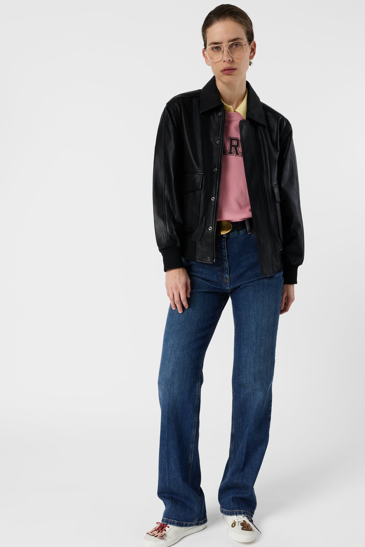 Person wearing a black leather jacket, pink shirt, blue jean brut in a coupe large style, white shoes, and glasses stands against a plain white background, looking at the camera with a neutral expression.