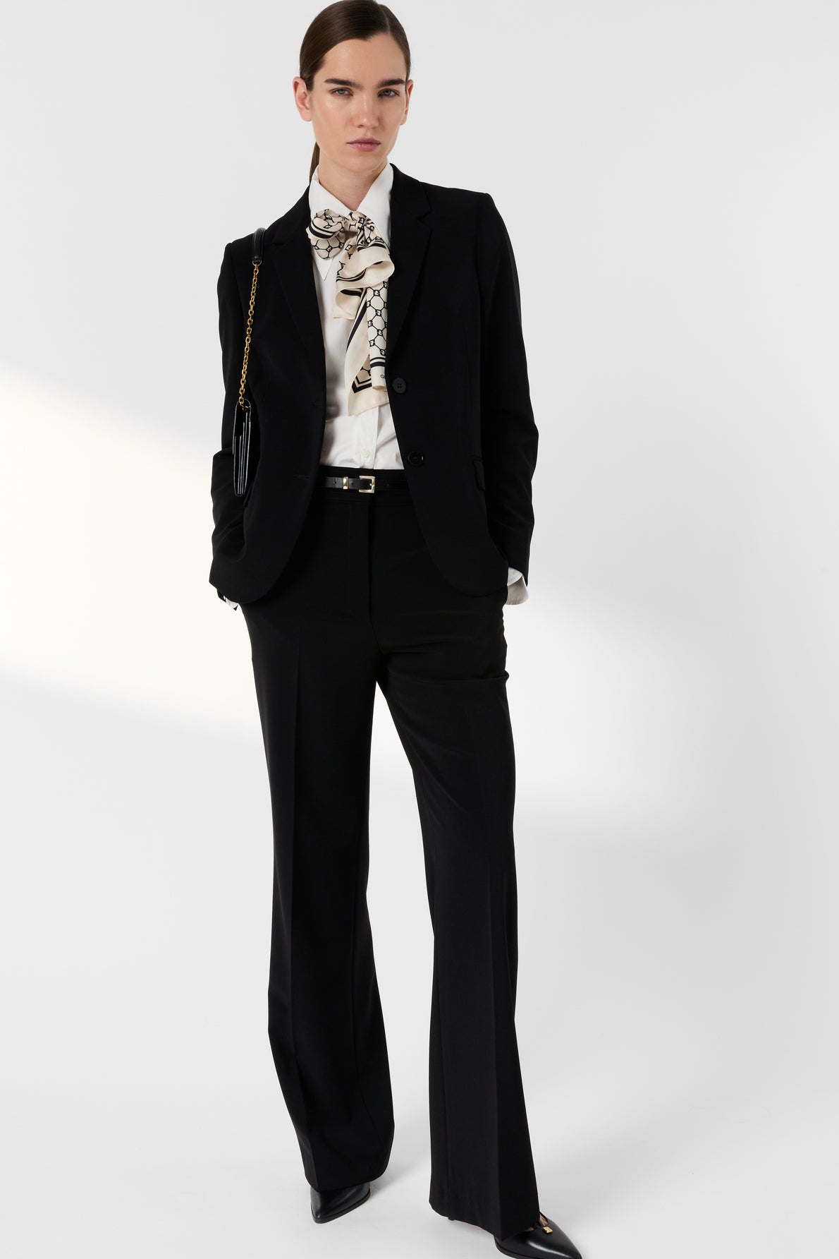 Woman wearing a black suit with chic wide-leg trousers (pantalon chic), a white blouse with a printed scarf tied at the neck, black shoes, and a small black shoulder bag, standing against a plain white background.