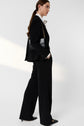 A person with long brown hair in a low ponytail wears a pantalon noir femme suit with a white shirt and black heels, carrying a black shoulder bag with a gold chain, standing against a plain light background.