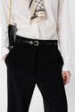 A person in a white blouse, pantalon fluide noir femme with a thin black belt featuring a gold buckle, holding a black purse with a gold chain strap, and wearing a patterned scarf.