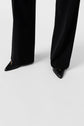 A person wearing a pantalon noir femme with a chic, wide-leg cut and black pointed-toe shoes stands on a light-colored floor. The pantalon fluide gracefully drapes over the shoes, partially covering them.