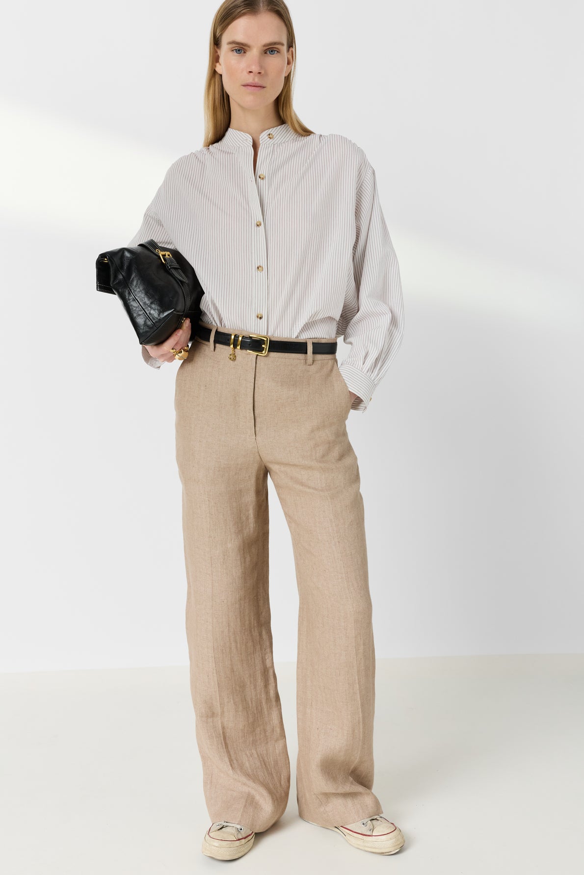 A person stands against a plain background, wearing a striped button-up shirt, beige wide-leg pants, white sneakers, and holding a black clutch bag under one arm.