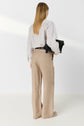 A person with straight, light brown hair stands facing away, wearing a striped long-sleeve blouse, beige wide-leg pants, and holding a black bag under one arm in a minimal, white room.