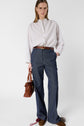 A woman wearing glasses, a white blouse, and a pantalon large femme in blue stands holding a brown handbag, looking at the camera against a plain white background. She completes her look with brown shoes.