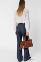 A woman with long blonde hair, wearing a striped shirt and a pantalon taille haute denim bleu foncé, stands facing away, holding a brown leather handbag in her right hand.