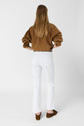 A woman with long hair, wearing a brown jacket, slim-fit jean blanc in coton stretch, and brown shoes, stands facing away against a plain white background.