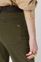 A person wearing olive green pants with a gold logo on the pocket, a beige shirt, a braided black belt, and several chunky gold rings on their hand.