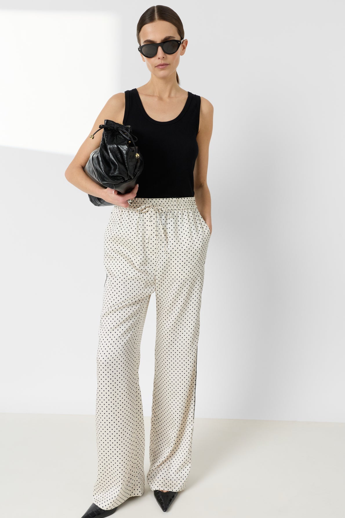 Woman wearing black sunglasses, a black sleeveless top, and white polka dot wide-leg pants stands against a plain background, holding a black handbag under her arm.