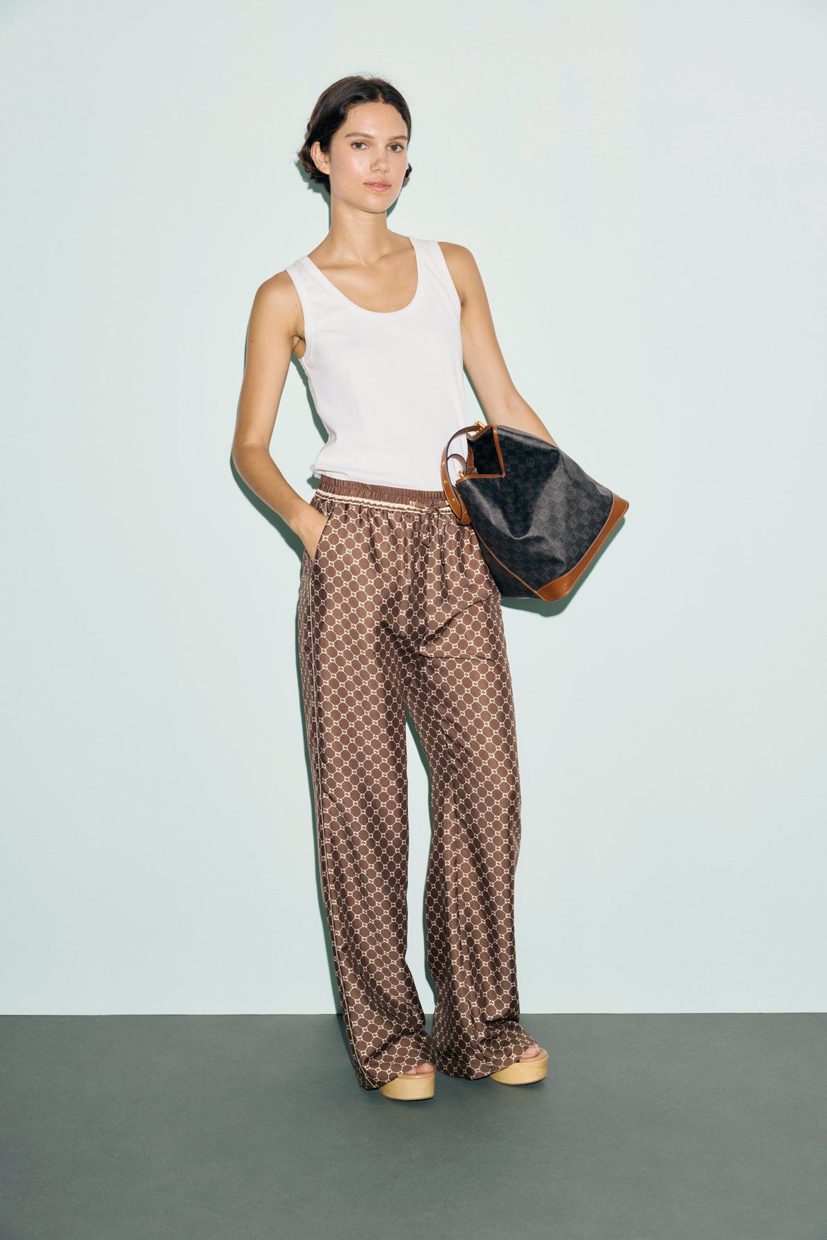 Woman stands against a pale background wearing a white sleeveless top, édition limitée patterned wide-leg pants, and sandals, holding a large black and brown monogramme tote bag.