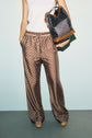 A person wearing a white sleeveless top and édition limitée patterned brown wide-leg pants with hands in pockets, holding a black and brown monogramme tote bag.