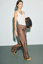 A woman walks wearing a white sleeveless top, édition limitée brown patterned wide-leg pants, platform sandals, and carrying a large patterned tote bag with a monogramme design against a plain light background.