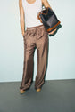 A person wearing patterned pantalon marron satin, a white sleeveless top, tan platform shoes, and holding a black and brown monogramme bag édition limitée against a plain background.