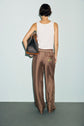 A person stands facing away, wearing a white tank top and a pantalon marron satin with an intricate pattern, holding a large black bag on their right shoulder. The background is light blue, and the person has short, dark hair. This effortlessly chic look exudes understated elegance, reminiscent of an édition limitée collection.