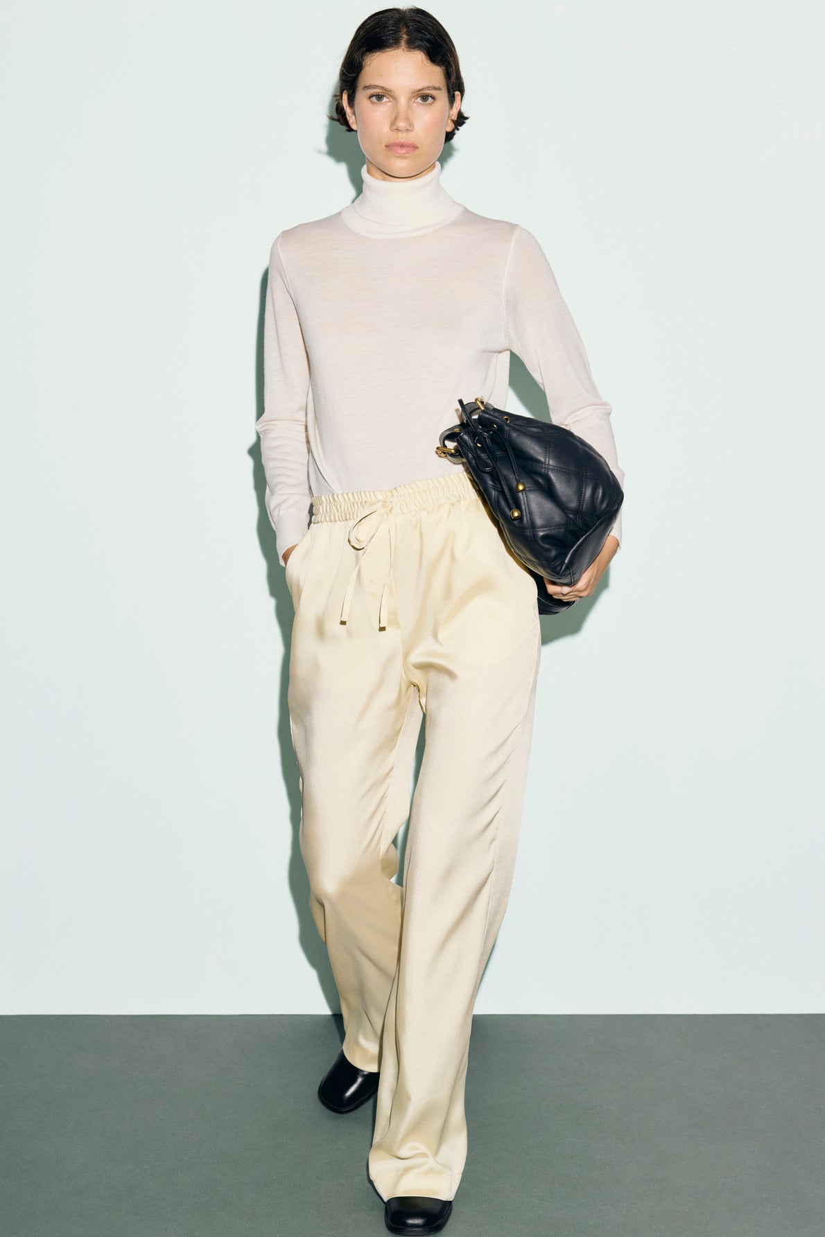 Woman wearing a sheer white turtleneck, cream pantalon satiné à taille élastiquée, and black shoes, holding a large black bag under her arm, standing against a plain light background.