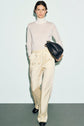 Woman wearing a sheer white turtleneck, cream pantalon satiné à taille élastiquée, and black shoes, holding a large black bag under her arm, standing against a plain light background.