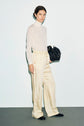 A woman wearing a white turtleneck and cream wide-leg trousers à taille élastiquée stands against a plain background, holding a black handbag and wearing black shoes.