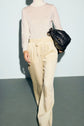 A person wearing a sheer white turtleneck and beige pantalon satiné with taille élastiquée holds a black clutch bag under one arm, walking against a plain light background.