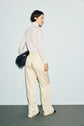 A person stands against a pale background, wearing a light turtleneck and cream wide-leg pantalon satiné with a taille élastiquée, paired with black shoes. They carry a black bag over one shoulder while looking back over their left shoulder.