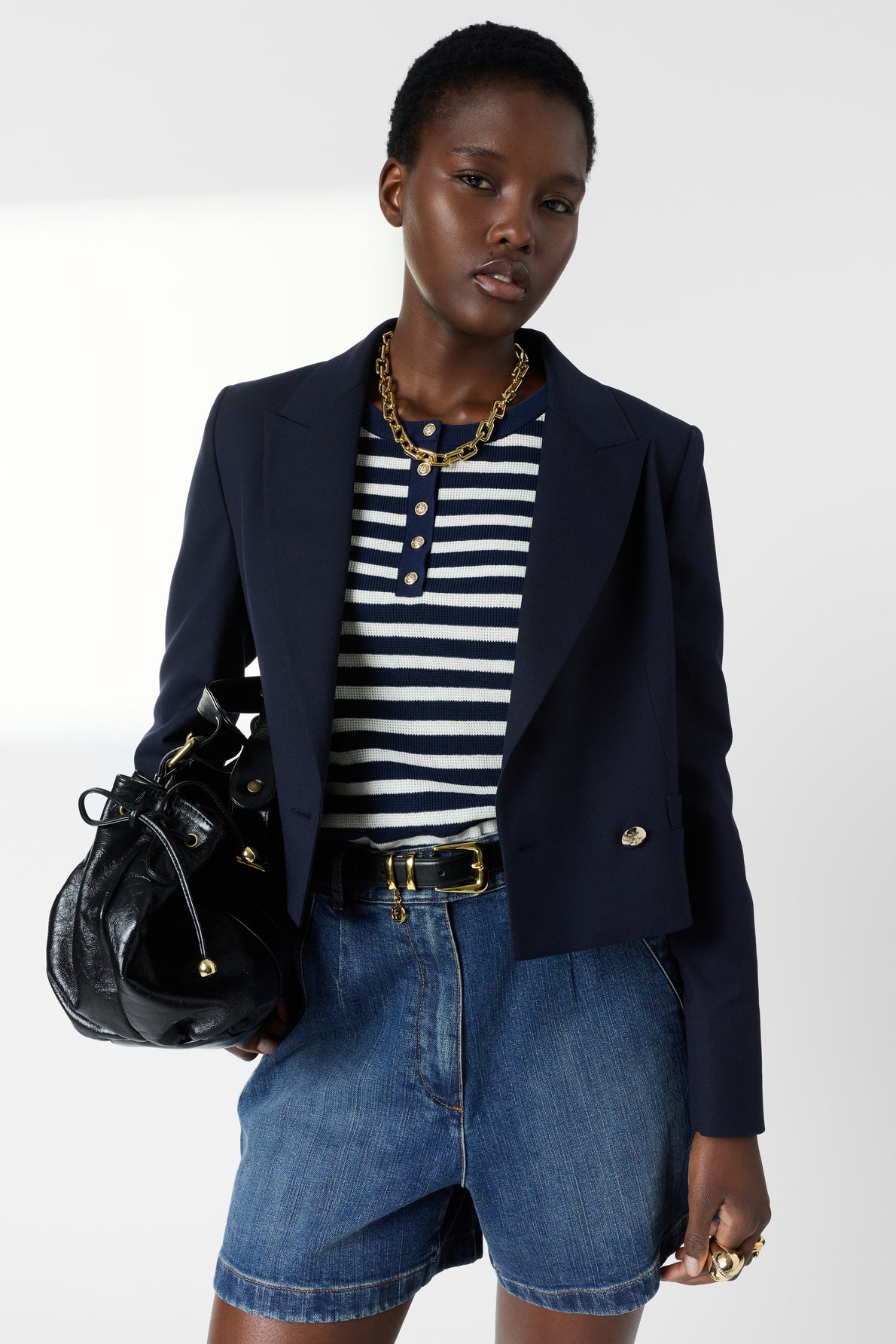 Woman wearing a navy blazer, striped shirt, denim shorts, and a chunky gold necklace, holding a black handbag, standing against a plain background.