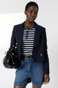 Woman wearing a navy blazer, striped shirt, denim shorts, and a chunky gold necklace, holding a black handbag, standing against a plain background.