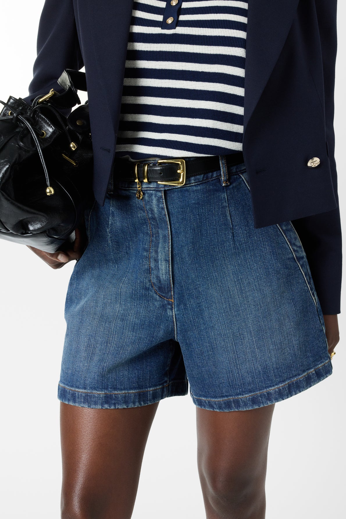 Person wearing a navy blazer, striped shirt, denim shorts, and a black belt, holding a shiny black handbag. Only midsection and part of the legs are visible against a plain background.