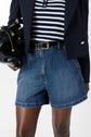 Person wearing a navy blazer, striped shirt, denim shorts, and a black belt, holding a shiny black handbag. Only midsection and part of the legs are visible against a plain background.