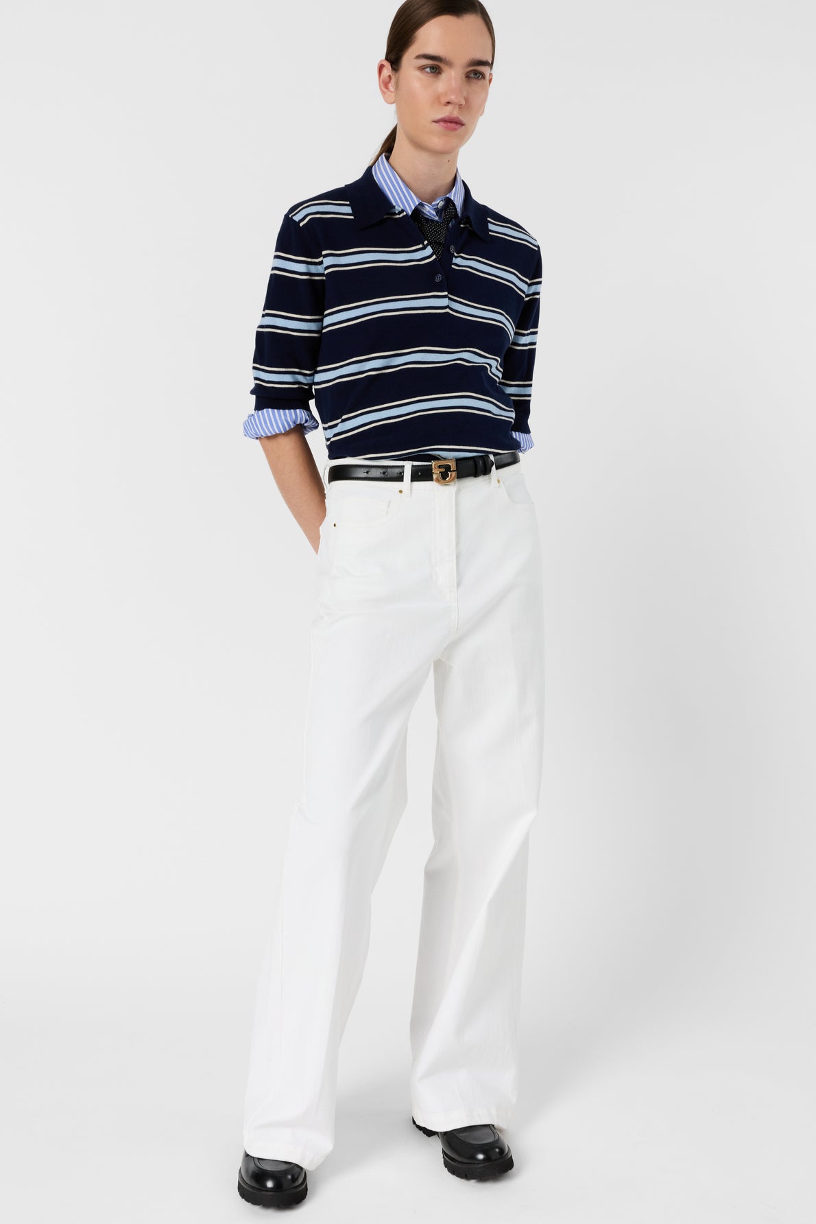 A person wearing a navy and white striped long-sleeve shirt tucked into a high-waisted pantalon long blanc à coupe large with a black belt, black shoes, and a blue collared shirt underneath, standing against a plain white background.