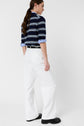 Person standing sideways, wearing a navy and white striped long-sleeve shirt, white wide-leg pantalon long, and black shoes. The shirt sleeves are partially rolled up, revealing a blue inner lining. The jean blanc adds a modern coupe large silhouette against the plain white background.