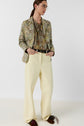 A person with glasses wears a floral jacket, patterned blouse, wide-leg cream pantalon femme, a black belt, and brown shoes, standing with hands in pockets against a plain white background.