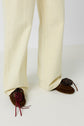 A person wearing loose, off-white pantalon femme and brown moccasins with decorative red and purple pom-poms stands on a white surface.