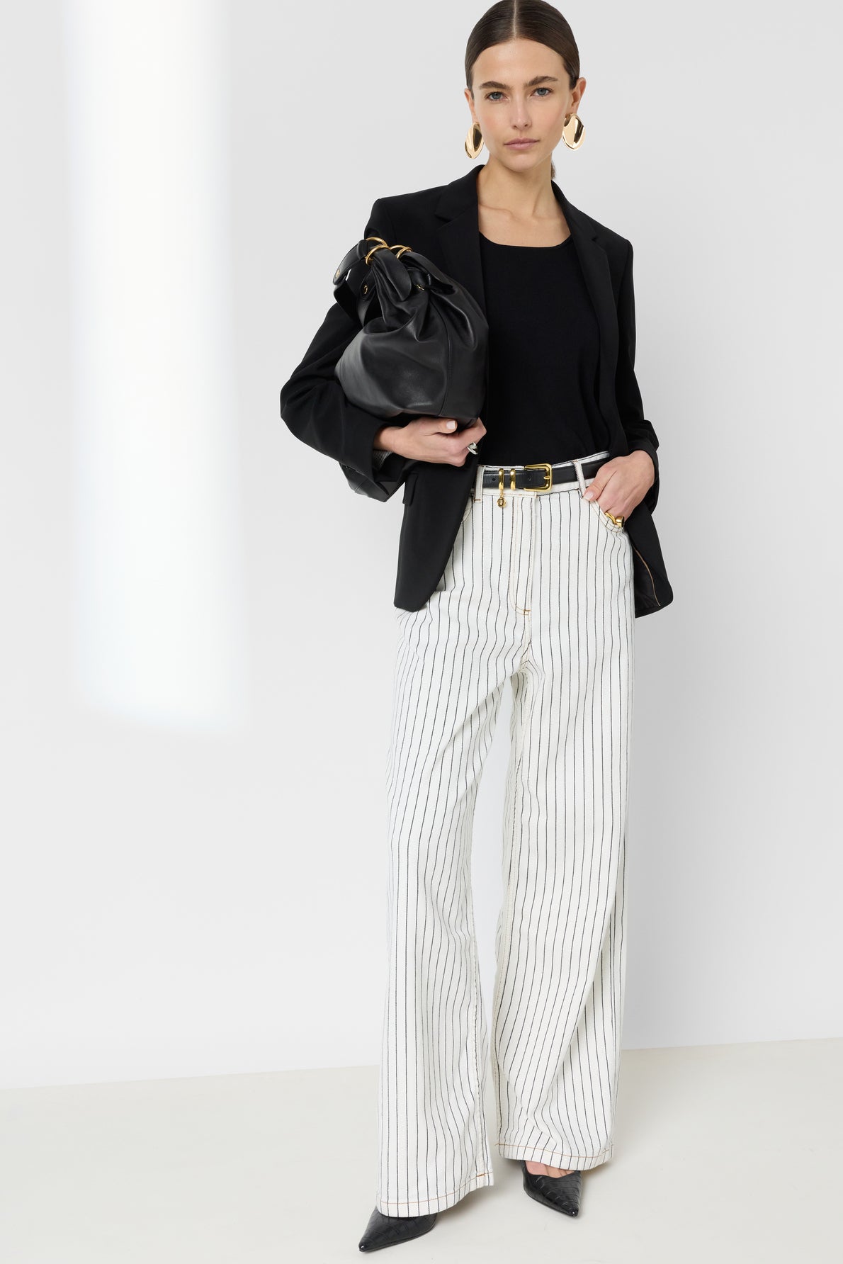 Woman in a black blazer, black top, and white high-waisted pinstripe pants reminiscent of a chic jean rayé holds a large black bag. She wears gold earrings and black pointed shoes, standing against a plain white background.