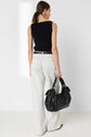 A woman from behind wears a black sleeveless top, white pinstriped wide-leg trousers reminiscent of a jean rayé style, and a black belt. She is carrying a large, soft black leather hobo bag with gold hardware in her right hand. She has a low ponytail and gold hoop earrings.