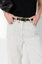 Person wearing a jean rayé—white pants with black vertical stripes—a black belt with a gold buckle, and a black sleeveless top, with hands resting in the front pockets.