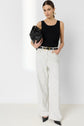Woman standing against a plain background, wearing a black sleeveless top, white high-waisted jean rayé pants, black pointed heels, holding a black handbag, with hair pulled back and gold hoop earrings.