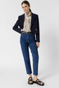 Woman stands against a plain background wearing blue jeans, a beige patterned blouse, a dark blazer, black belt, and black sandals. She has straight brown hair pulled back and a neutral expression.