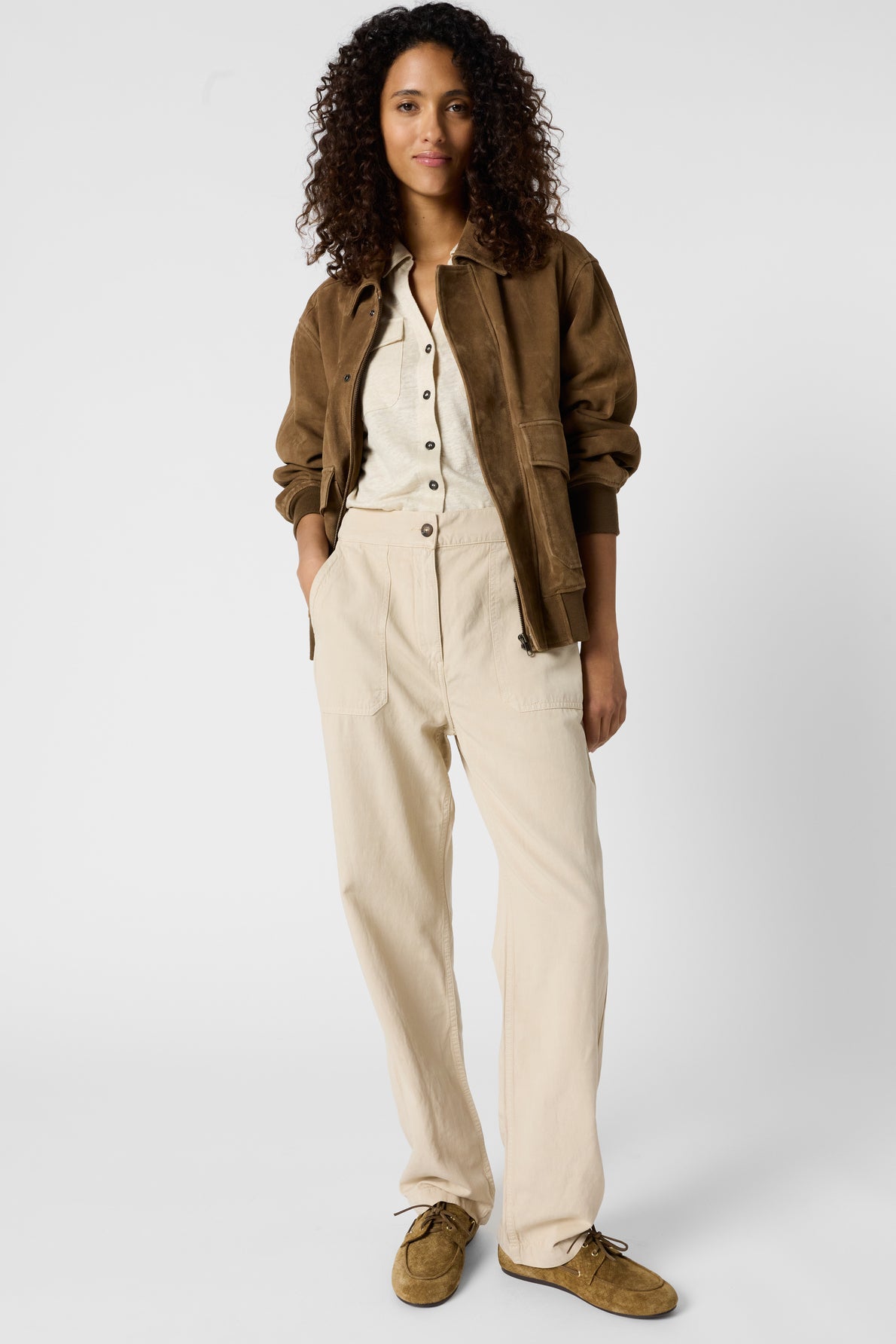 A person with curly hair wears a brown jacket over a cream button-up shirt and beige pantalon lin, standing with hands in pockets against a plain white background.