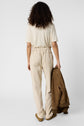 A person with curly hair, seen from behind, wears a beige t-shirt and light-colored pantalon 7/8, holding a brown jacket in their right hand against a plain white background.