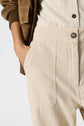 A person wearing light beige high-waisted pantalon lin with grandes poches and button detail, a cream shirt, and a brown jacket, with one hand in the pocket. Only the torso and upper legs are visible.