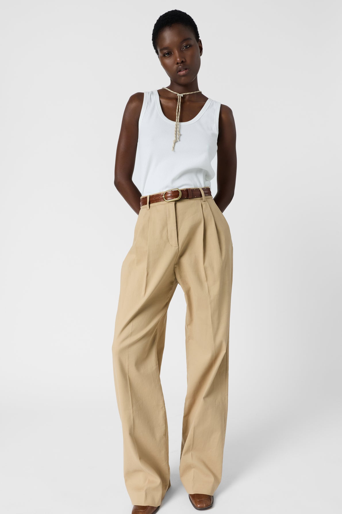 A person stands against a white background wearing a white sleeveless top, tan high-waisted pleated trousers, a brown belt, brown shoes, and a beaded necklace. Their hands are in their pockets.