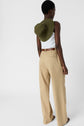 A person stands facing away, wearing a white sleeveless top, beige wide-leg pants with a brown belt, and holding an olive green hat over their shoulder. The background is plain white.