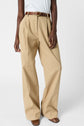 Person wearing high-waisted, wide-leg beige trousers with pleats, a brown belt, white sleeveless top, and brown square-toe shoes, standing against a plain light background.