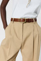 A person wearing beige pleated trousers with hands in pockets, a brown leather belt with a gold buckle, and a white ribbed top.