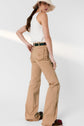 Woman in a white sleeveless top, beige jean ANNA à taille haute et coupe flare, woven belt, and wide-brimmed hat poses with one hand on her hip, facing backward. Her long hair is loose and she wears brown shoes with beaded details.