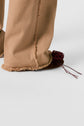 Person wearing wide-legged tan pants with frayed hems in a coupe flare style and flat brown shoes that have a dark red woven detail and long laces on a light background.